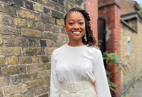 Nadia Odunayo, a black woman who is smiling and wearing a white top, standing in front of a brick wall.