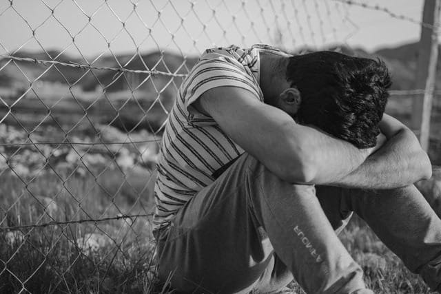 Black and white photo of man sitting on ground beside wired fence with his head on his arms