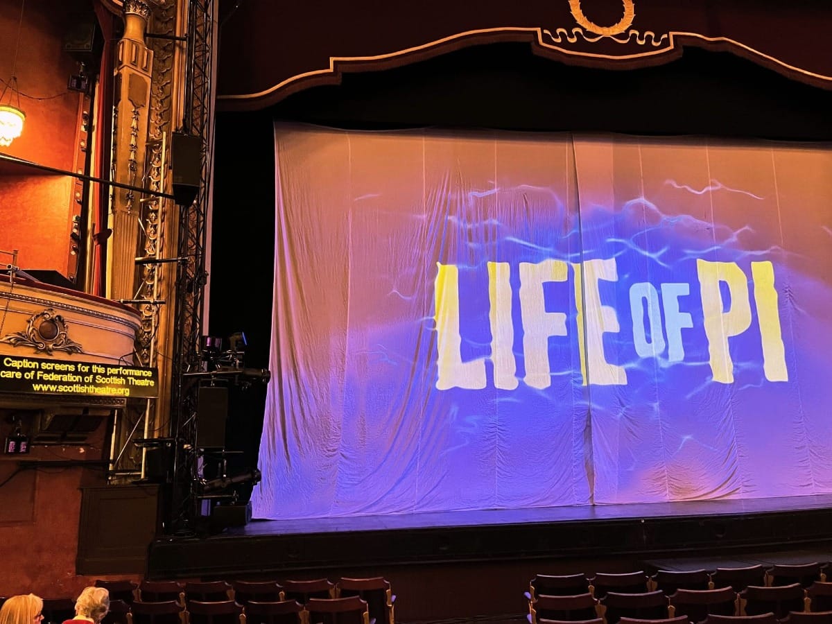 A theatre stage with a large canopy that has the sea with 'Life of Pi' written on top of it. On the left hand side, there is a captioning screen. Typed on the screen is "Caption screens for this performance, care of Federation of Scottish Theatre at www.ScottishTheatre.org".