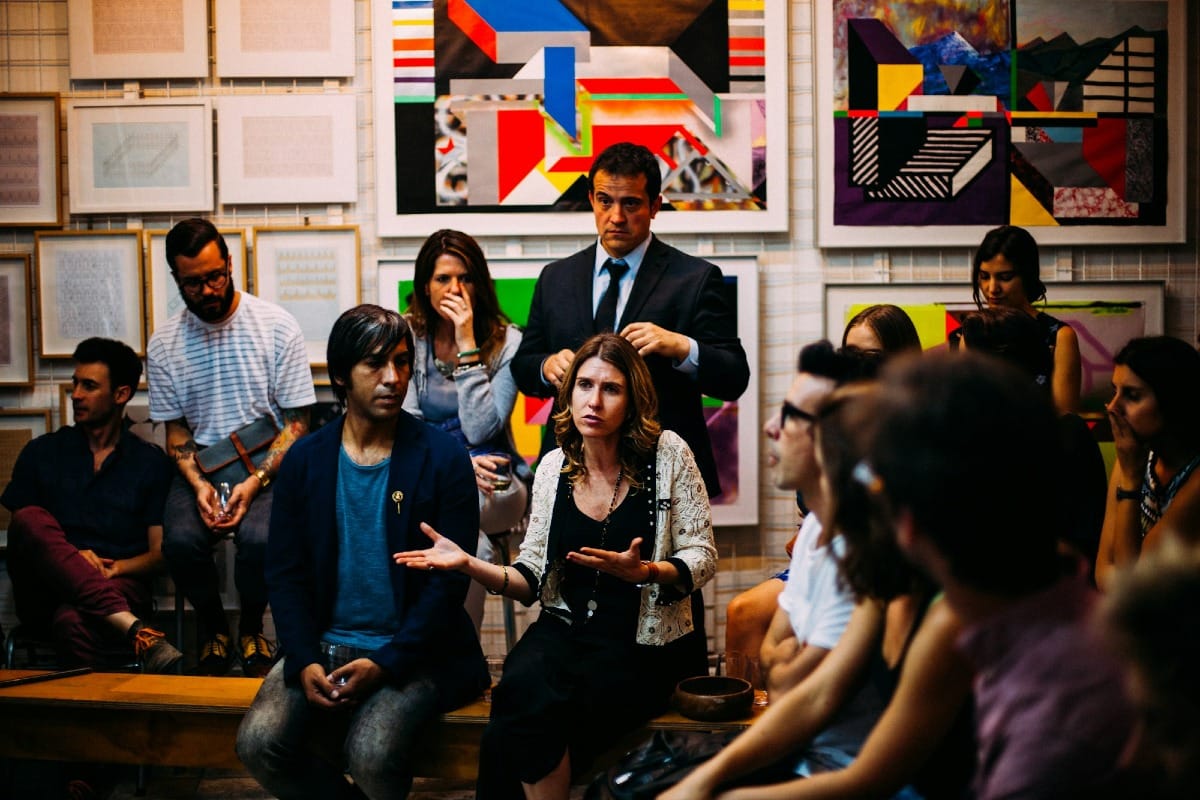 A group of people in a room facing one direction. One person is speaking to another person off-camera while everyone else is listening to the conversation.