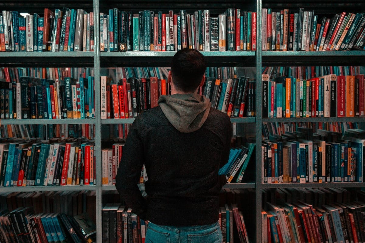 A man wearing black and grey jacket, standing in front of a large bookshelf