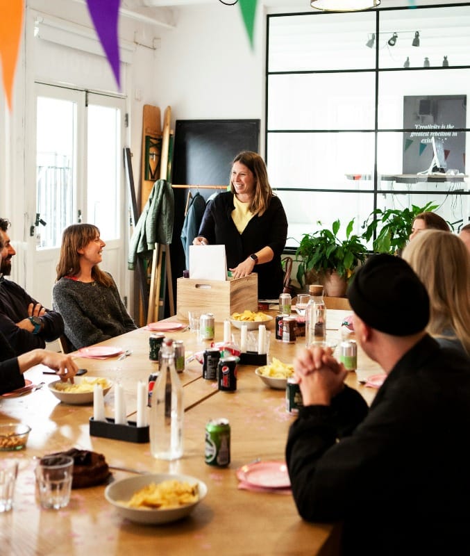 People in sitting around rectangular wooden table with drinks, snacks and appears to be in a social mood.