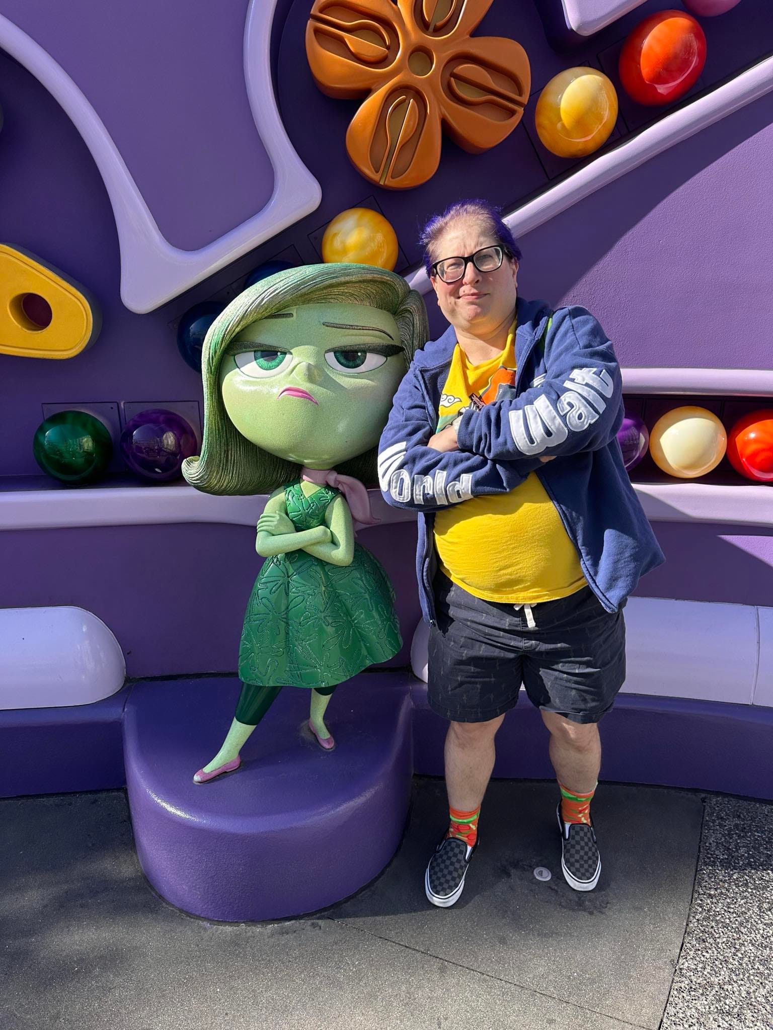 A person with purple hair, glasses, and a blue Disney World hoodie poses with a statue of Disgust from the Inside Out movies, mimicking the character's expression.
