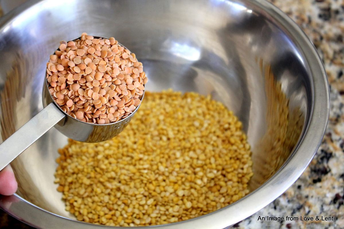 Dried red lentils in a metal scoop, being held above a metal bowl with more lentils in it.