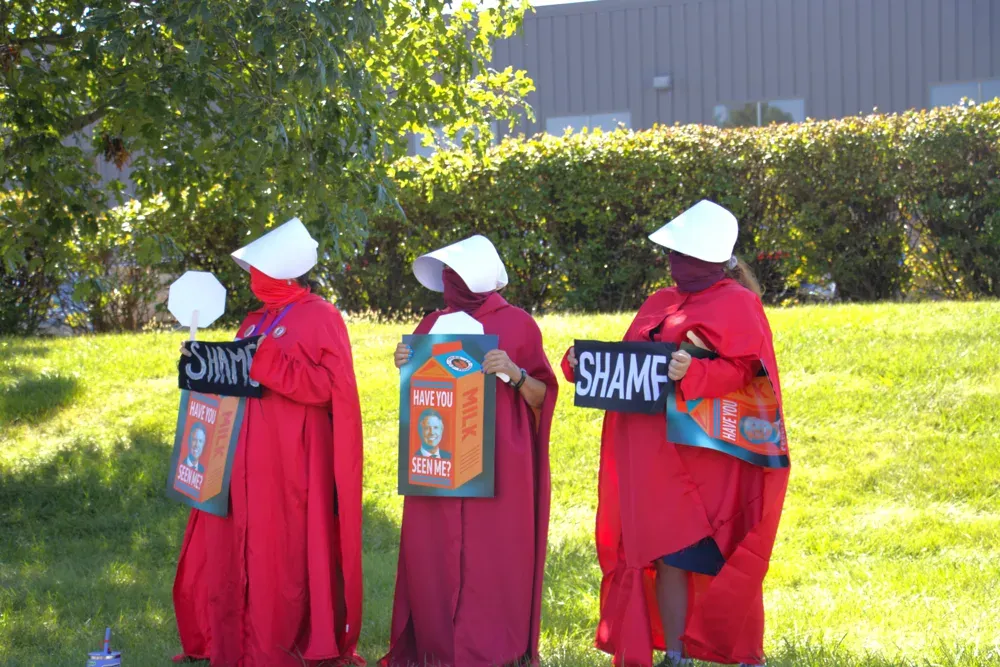 Protesters dressed as handmaidens hold anti-Roger Marshall signs outside a rally featuring Vice President JD Vance on Sept. 14, 2026. The signs reference allegations that Roger Marshall does not live in Kansas, a claim he has repeatedly denied. (Matthew Petillo/Citizen Journal)
