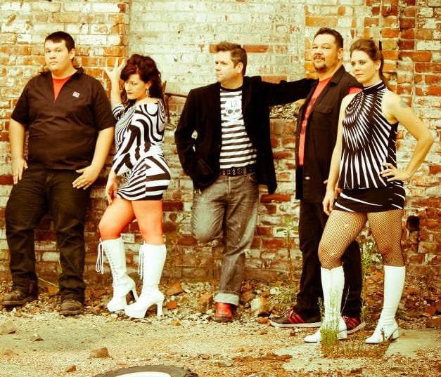 Members of a band, two women and three men standing in front of a brick wall