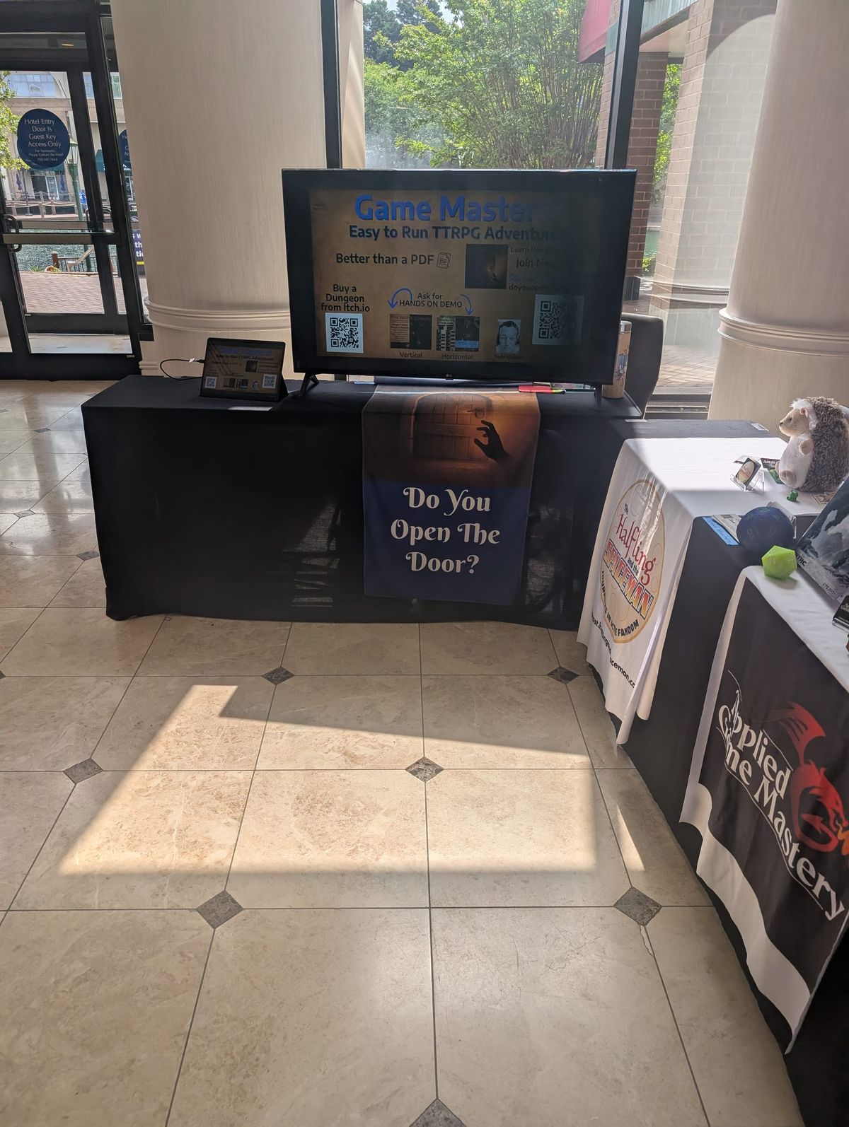 presentation table in hotel hall for Do You Open The Door? with tablet and TV display showing title screen for Game Masters