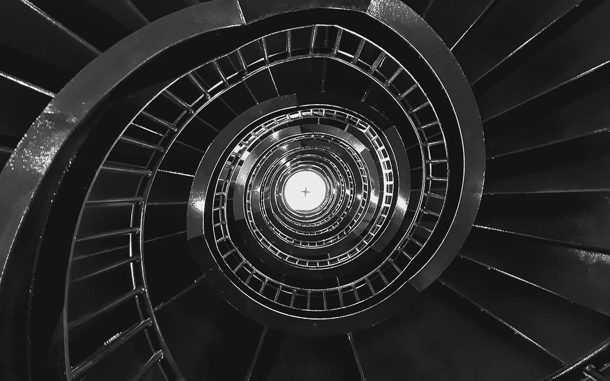 A spiral staircase from above, black and white, its curves tightening toward a point of light at the center. A mind circling the story of itself, always descending, never arriving.