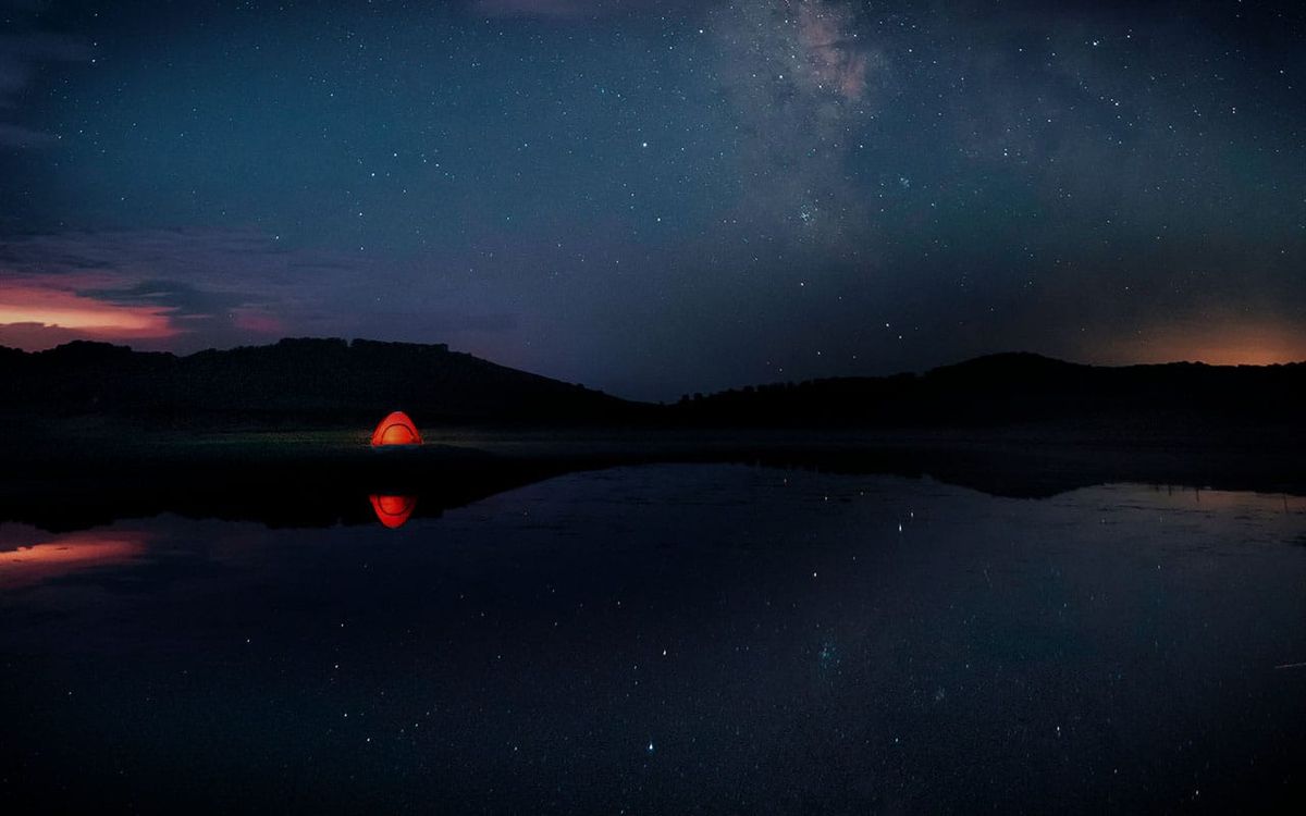 Starry night sky with the Milky Way reflected in still water, a small orange tent glowing at the shoreline.