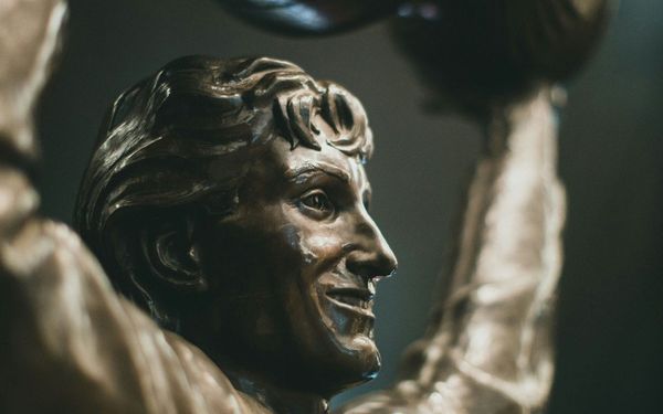 Dramatic close-up of an ancient Greek-style sculpture: a powerful figure mid-lift with a overflowing bucket, captured in stark monochrome.