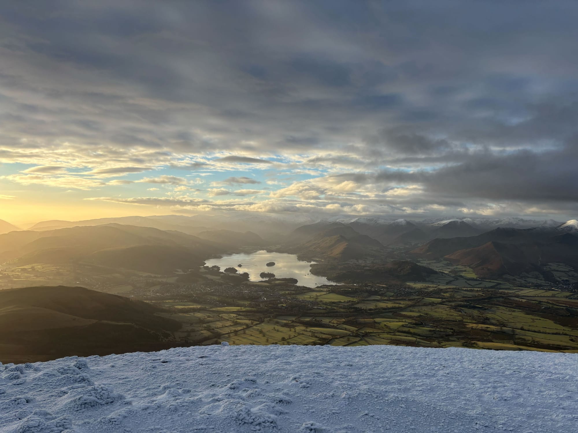 Sun low and off to the left, casting orange huges over a lake and other mountains in the background, with a green valley floor and snow on the floor in the foreground.