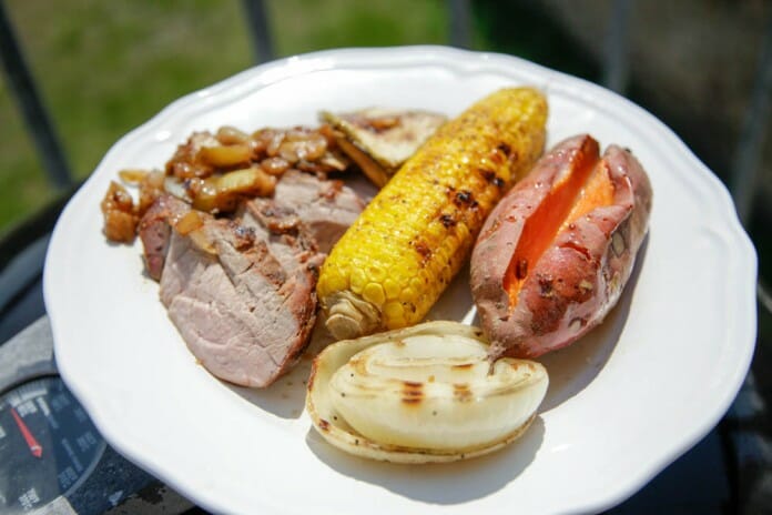 assiette blanche contenant des tranches de filet de porc fumé, un épis de maïs grillé, une patate douce et une grosse tranche d'oignon grillé
