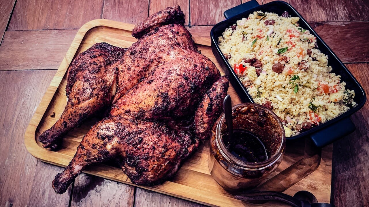 Un poulet en entier rôti sur une planche, un pot d'harissa et du taboulé.