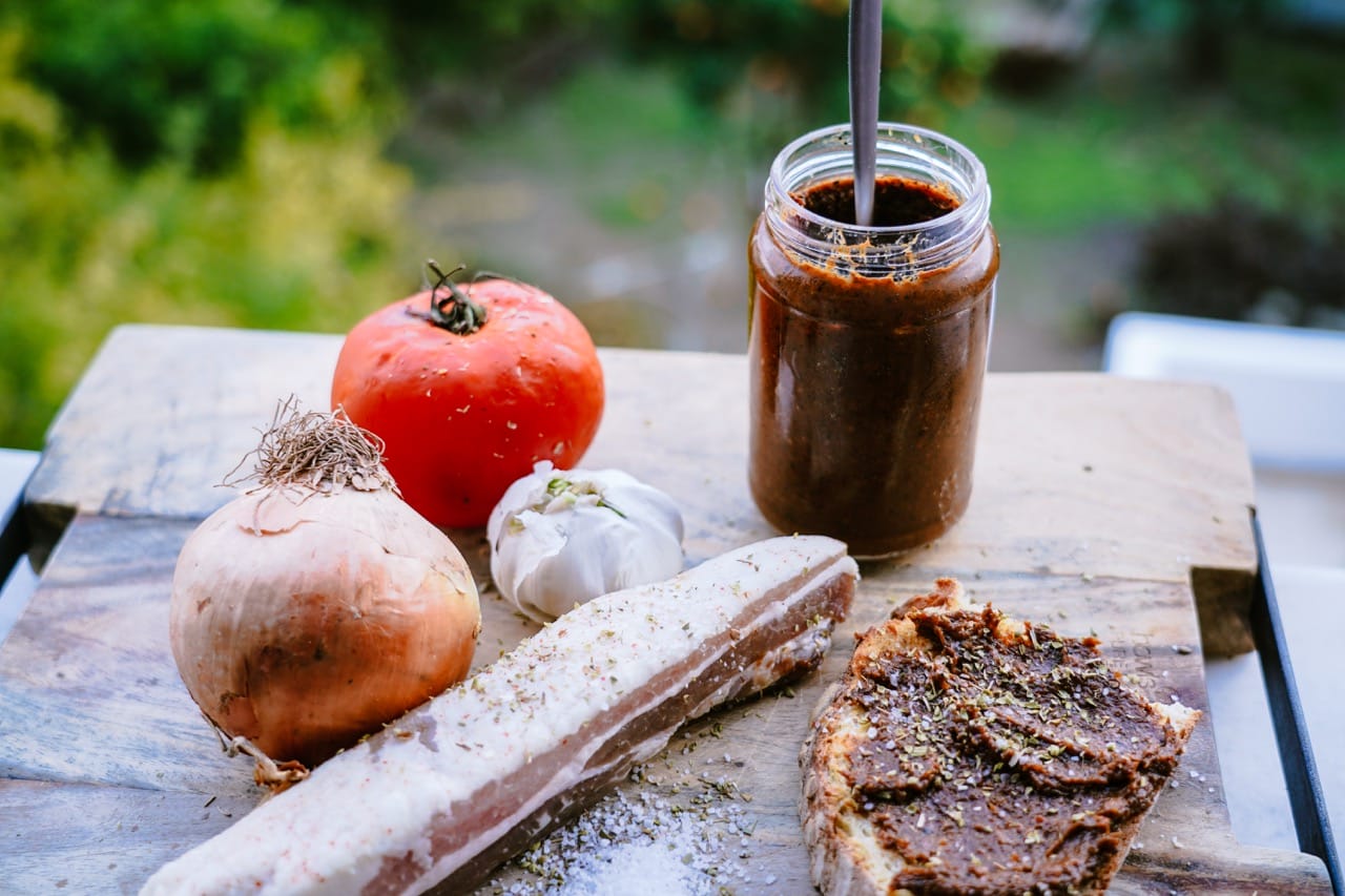 un pot de confiture de tomates, un bloc de bacon, une tartine avec de la confiture dessus, une tomate, un oignon et une tête d'ail