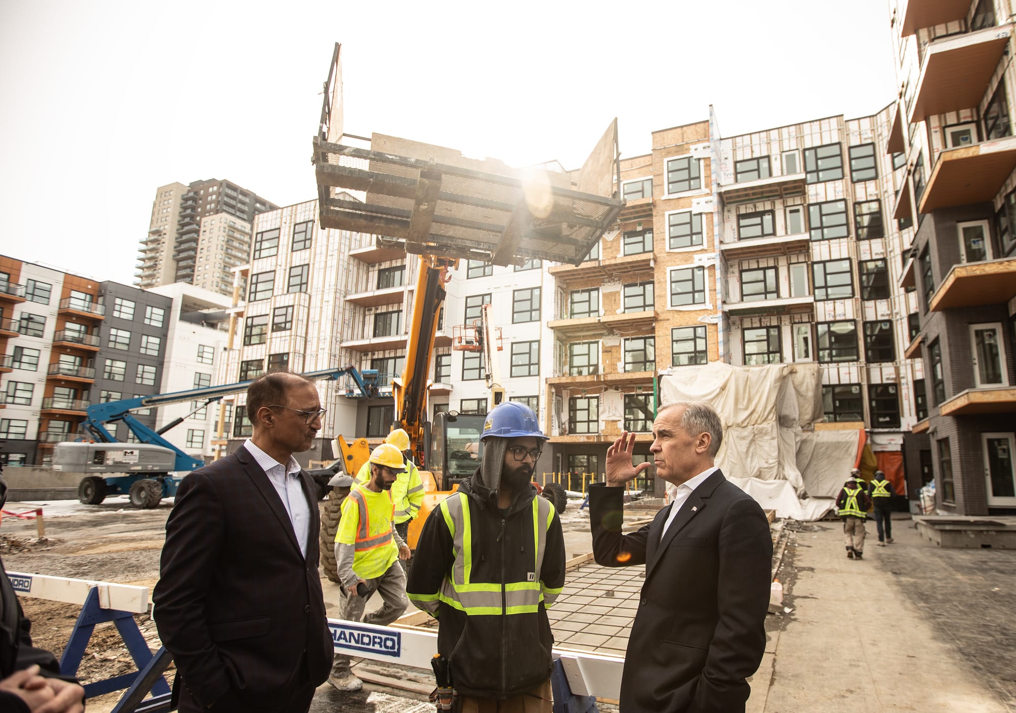 Amarjeet Sohi stands with Mark Carney and a construction worker in front of an in-construction apartment building.