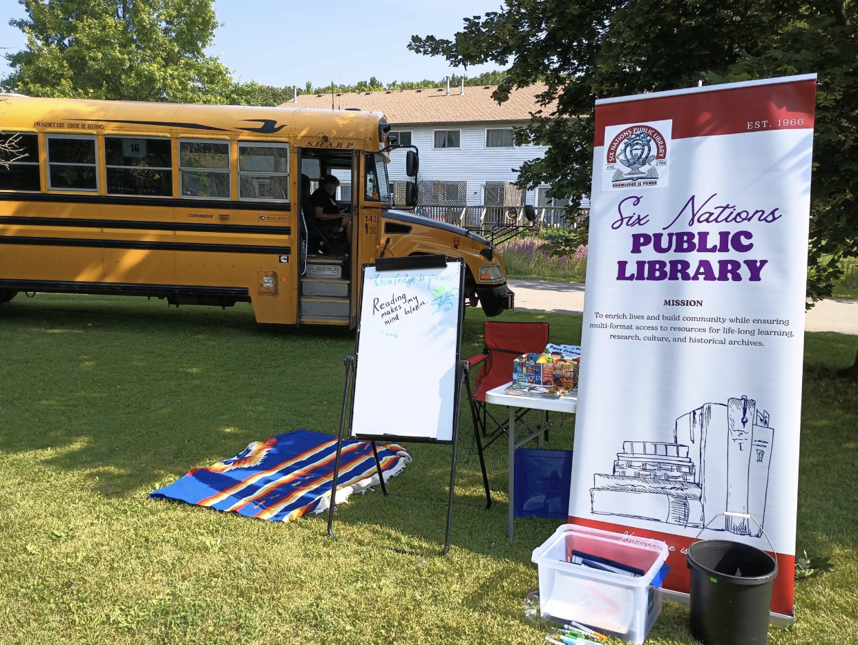 A sign for the Six Nations Public Library is seen on a lawn in front of a school bus.