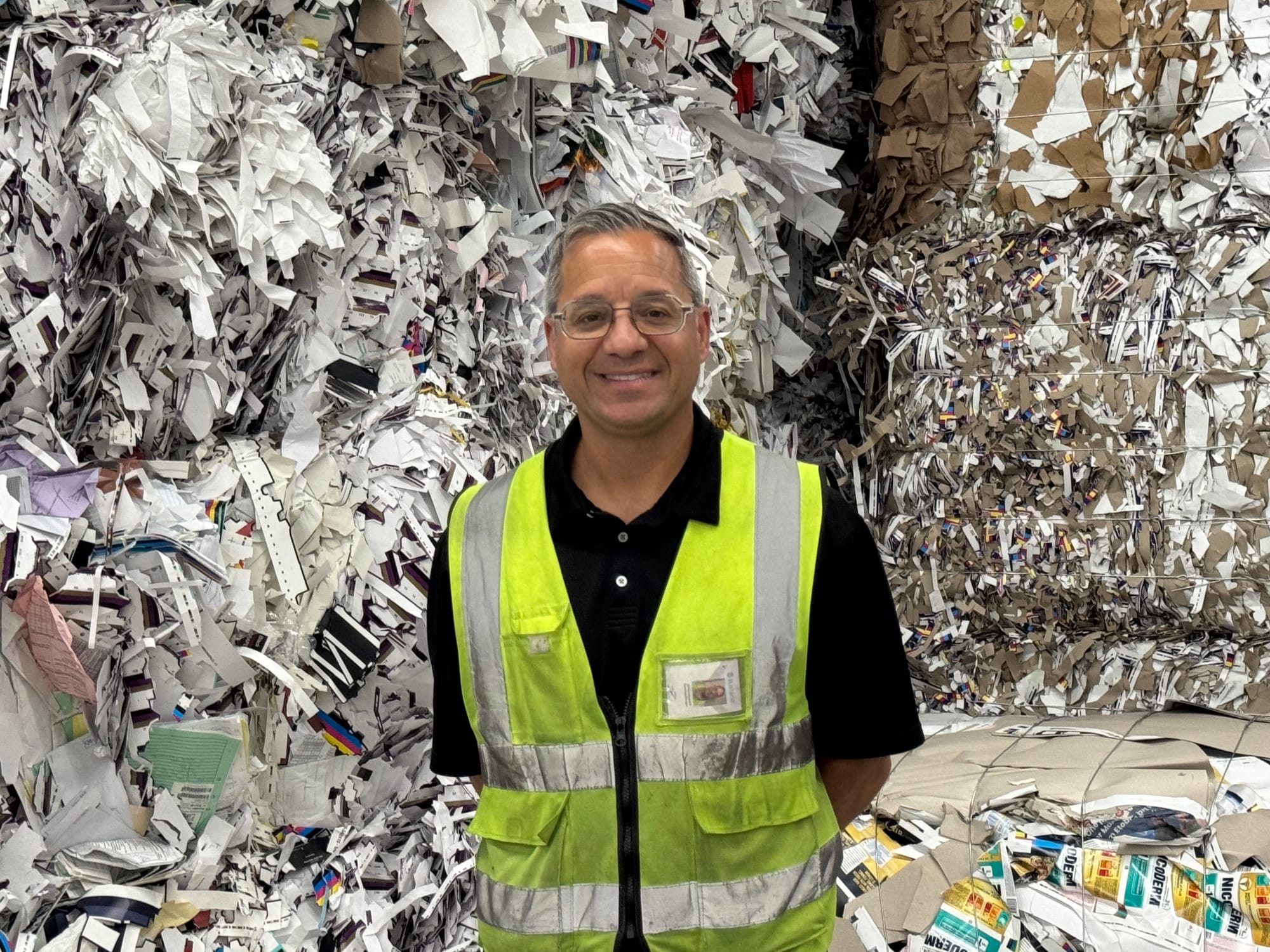 Sustana's corporate operations manager, David Campagnolo, stands in front of a wall of bound recycled paper wearing a yellow safety vest overtop of a black golf shirt. He wears glasses.
