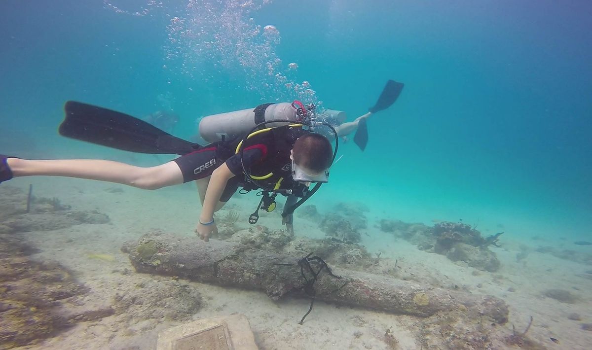 Buzo nada sobre un cañón antiguo cubierto de coral en el fondo arenoso de un museo vivo del mar