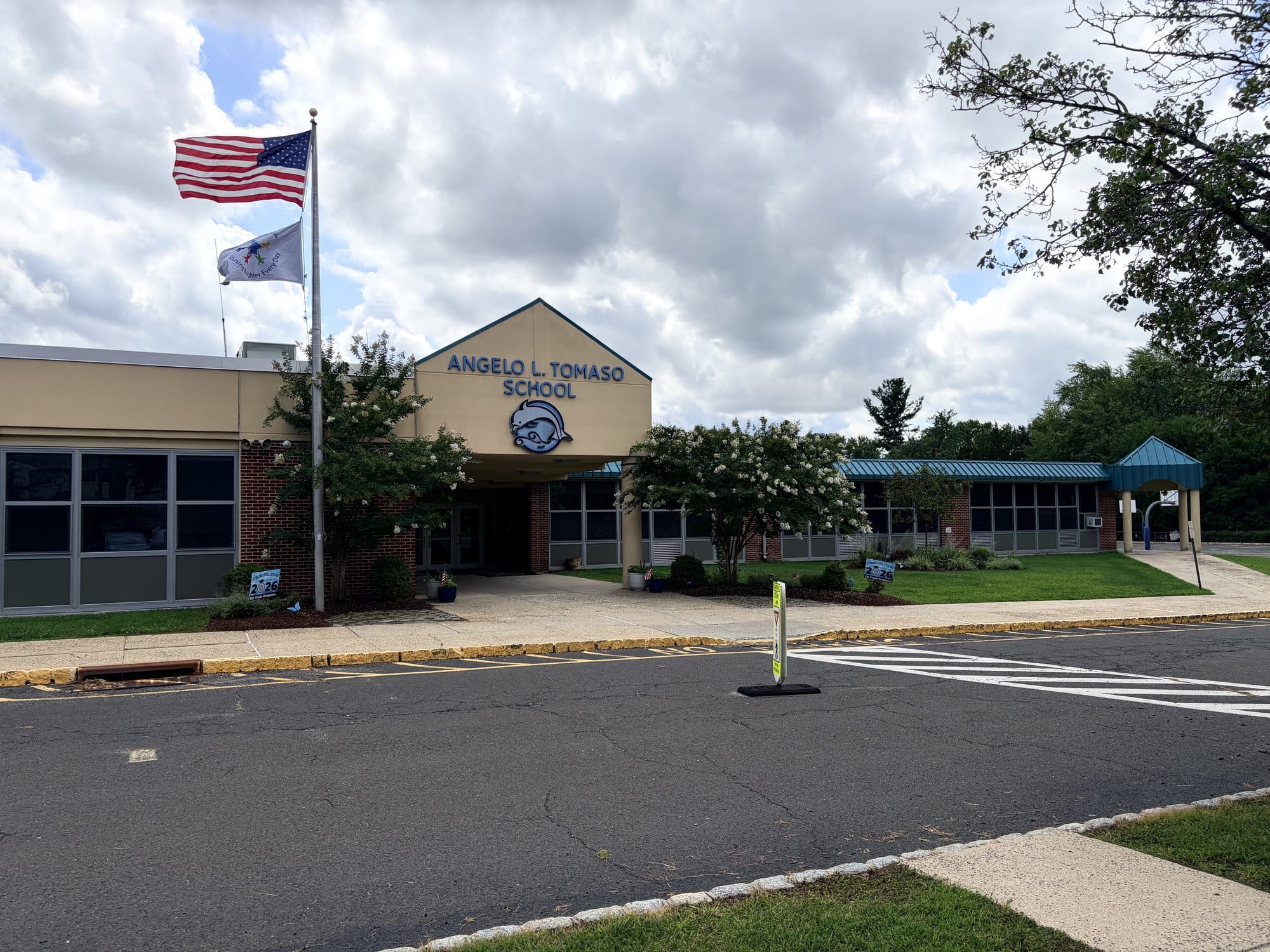 Angelo L. Tomaso School in Warren Township, a low tan and brick elementary building with ANGELO L. TOMASO SCHOOL lettered over the entrance beside a ram mascot, the American flag and a school flag on a pole out front.