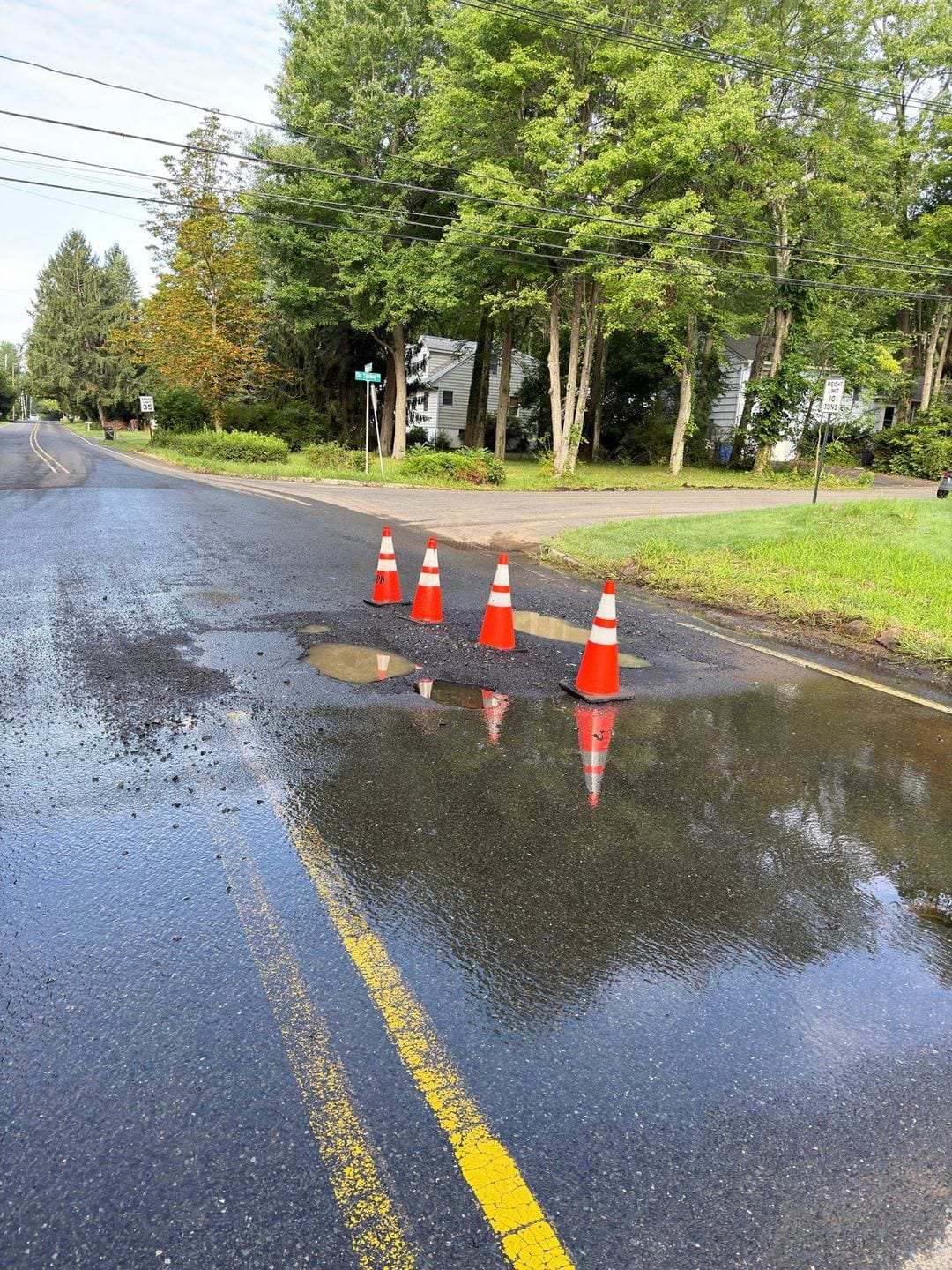 Water pooling across Reinman Road in the foreground, with four cones around the patched main cut at the Old Stirling Road corner.