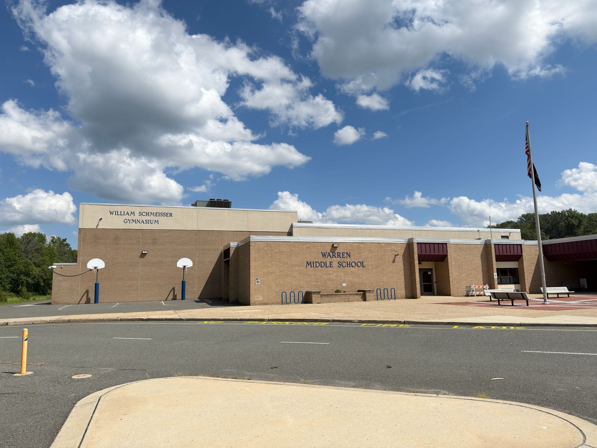 Warren Middle School, a low tan brick building with WARREN MIDDLE SCHOOL lettered beside the entrance and WILLIAM SCHMEISSER GYMNASIUM on the wing to the left, a flag pole out front.