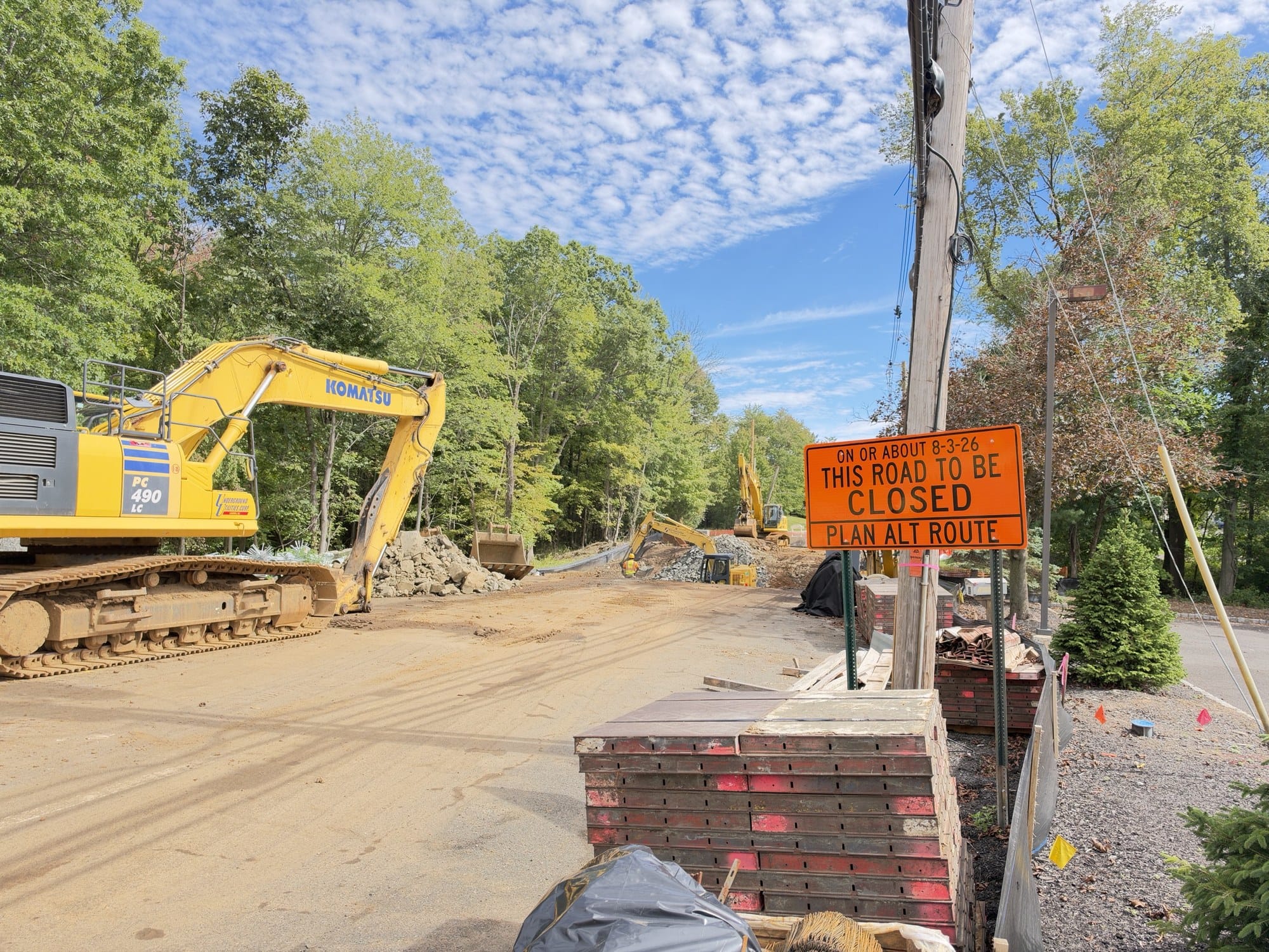 Stirling Road torn up for the bridge replacement, a yellow Komatsu excavator and rock piles at left, stacked concrete forms and an orange road closed sign at right.