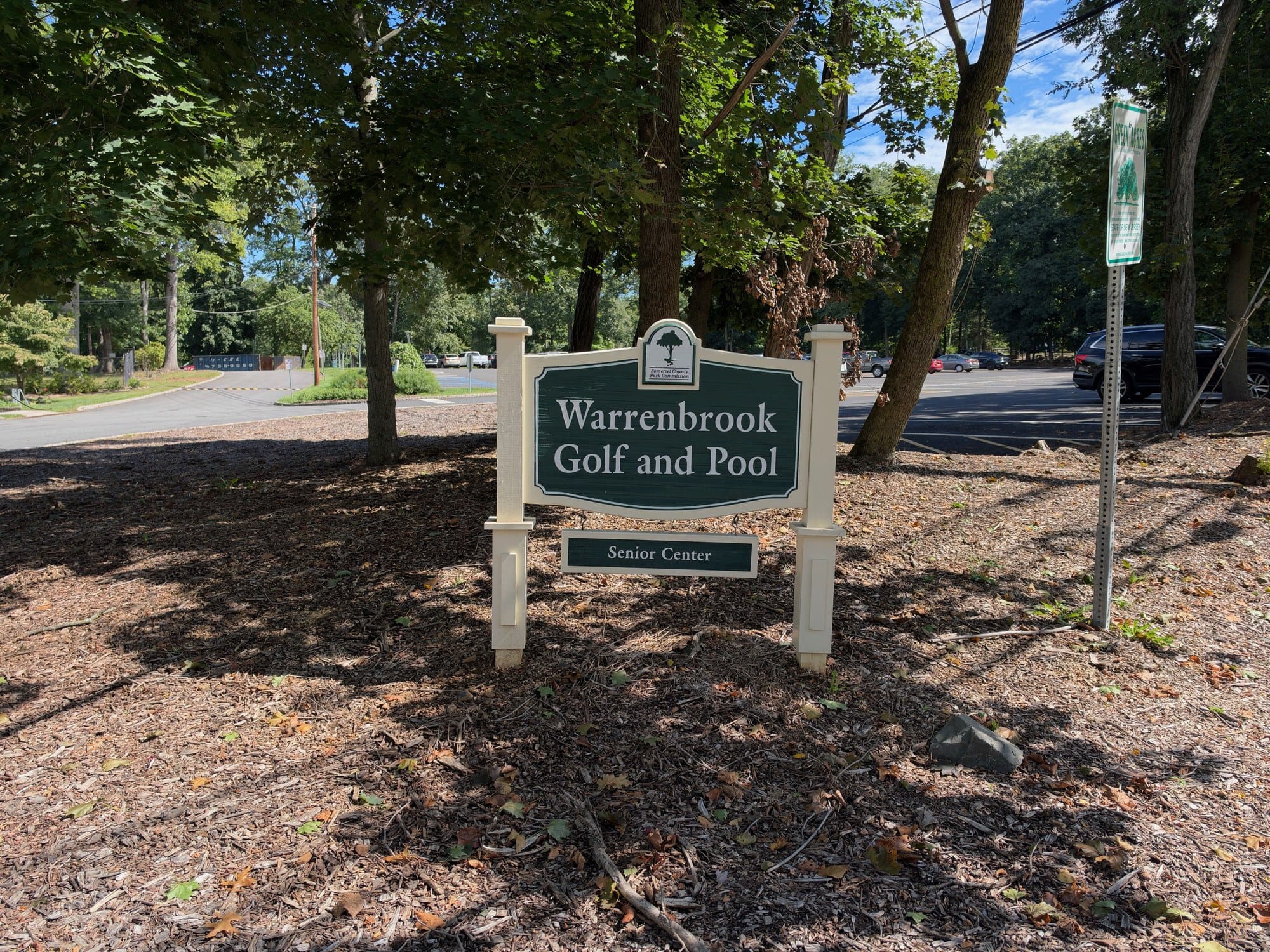 Warrenbrook Golf and Pool entrance sign with a Senior Center placard below, set among trees beside the parking lot at the Warrenville Road park entrance on a sunny late-summer day.
