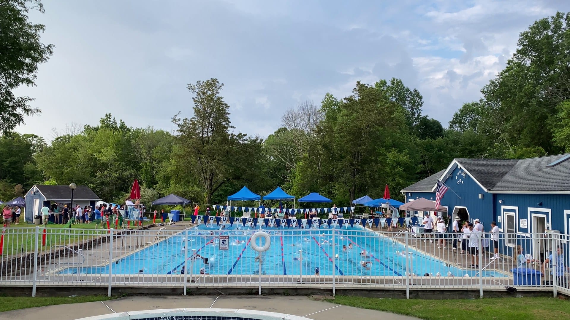 Outdoor pool with lane lines and a string of pennant flags reading Round Top Gators, blue canopy tents on the deck, and a blue clubhouse with an American flag.