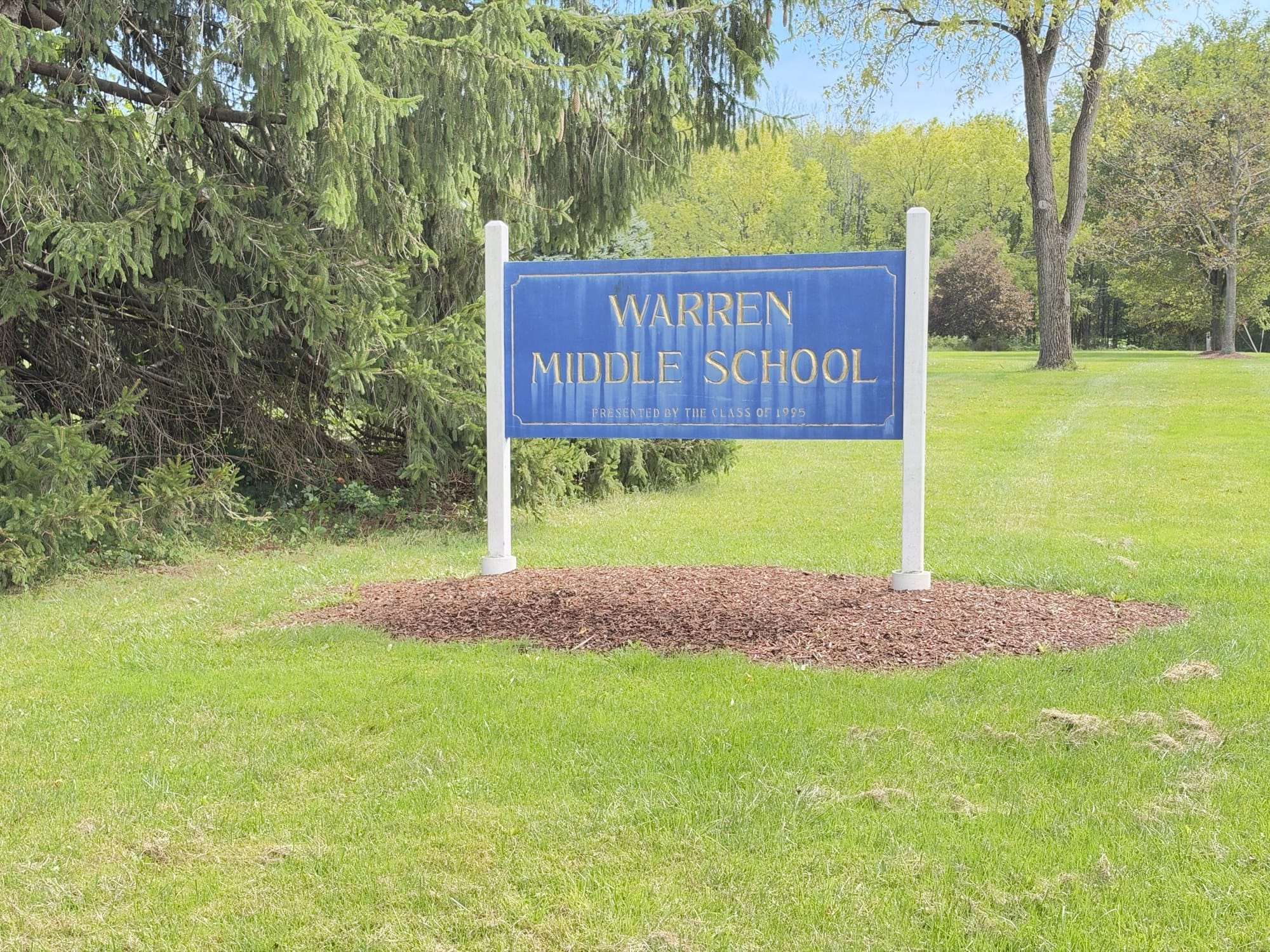 The Warren Middle School sign, blue with gold lettering and a line reading Presented by the Class of 1995, on white posts in a lawn beside a spruce.