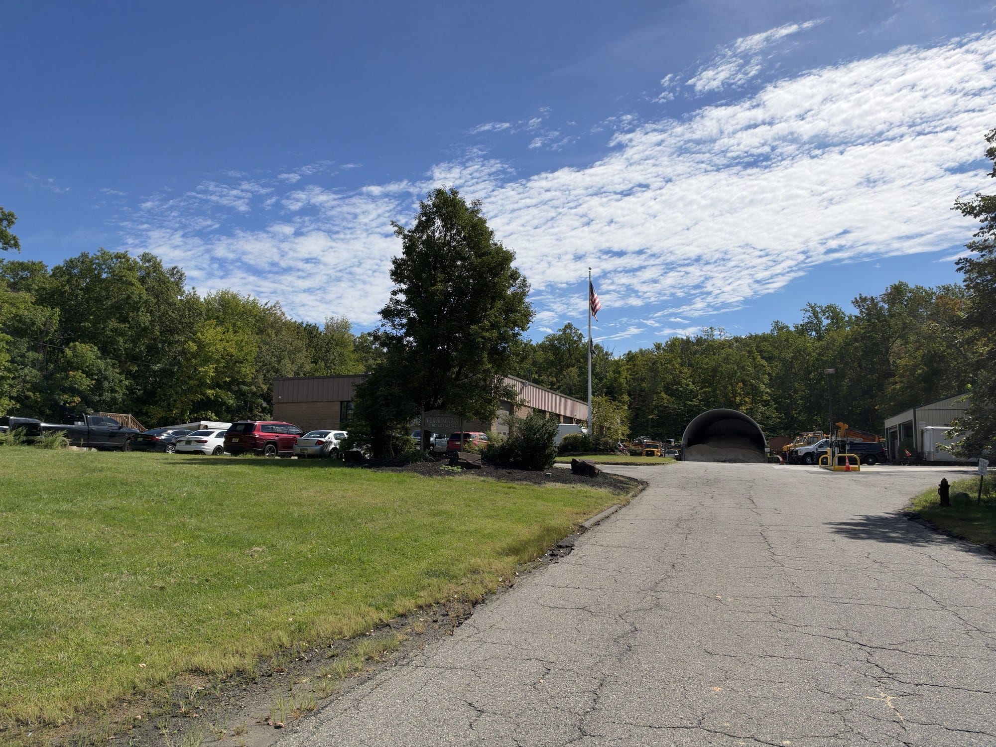The Warren Public Works yard, a tan brick building with a New Jersey's Finest Public Works Department sign, a flagpole, a domed storage shed and parked trucks at the end of a drive.