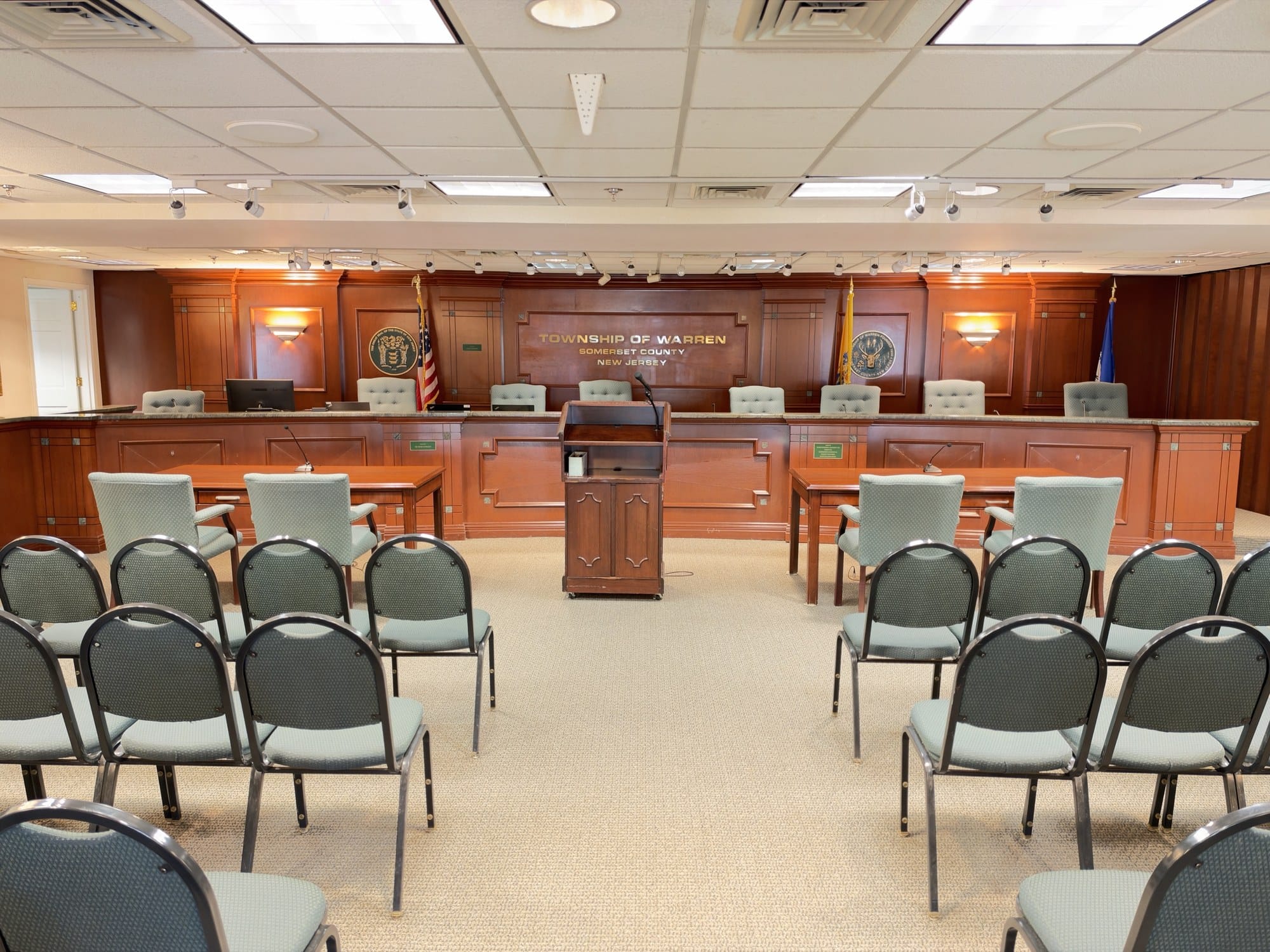 The Township of Warren dais, a wood paneled bench with the state and township seals and flags, empty chairs, a lectern and rows of public seating.