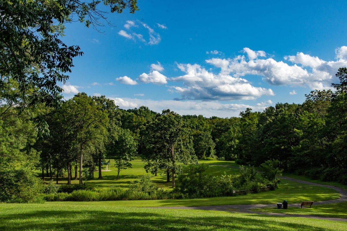 East County Park in Warren Township, an open mown lawn falling away to a tree line, a paved path curving past a bench on the right and a small pavilion in the distance.