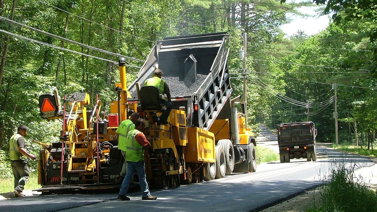 A road crew laying fresh asphalt behind a paver on a tree lined two lane road.