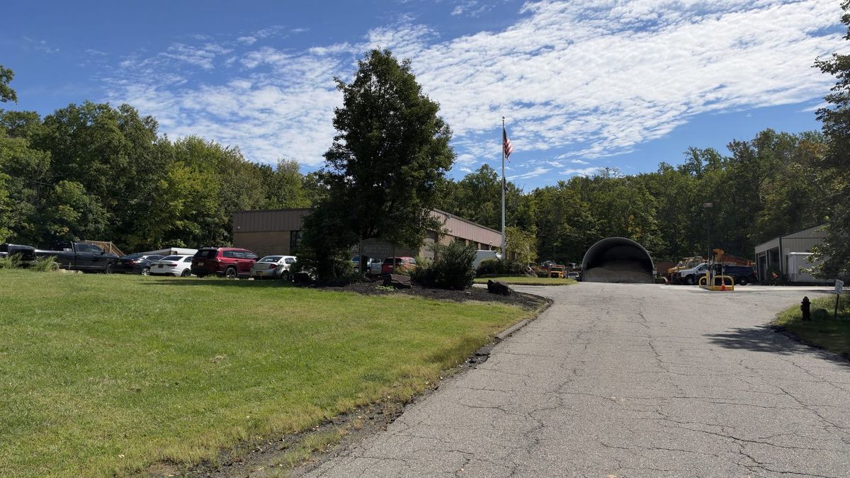 The Warren Public Works yard, a tan brick building with a New Jersey's Finest Public Works Department sign, a flagpole, a domed storage shed and parked trucks at the end of a drive.