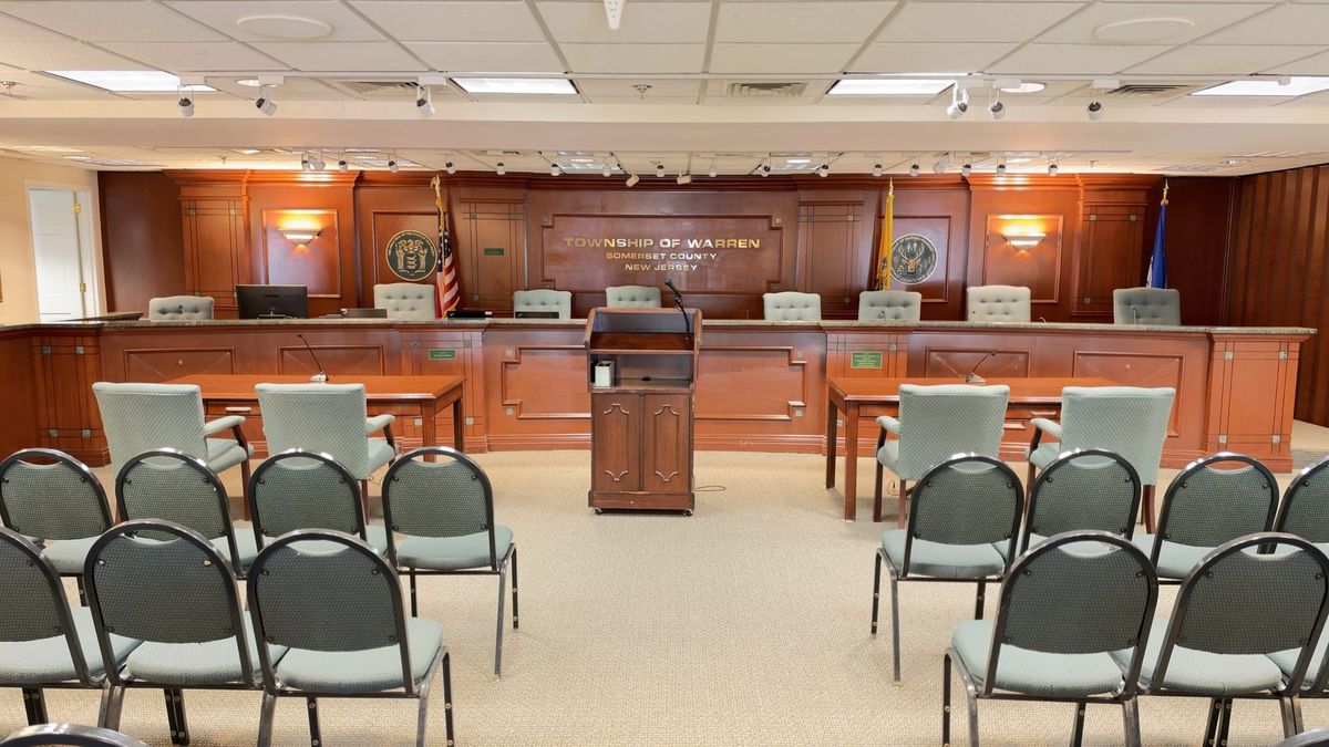 The Township of Warren dais, a wood paneled bench with the state and township seals and flags, empty chairs, a lectern and rows of public seating.