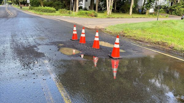 Four traffic cones around a patched water main cut on Reinman Road, water sheeting across the road at Old Stirling Road