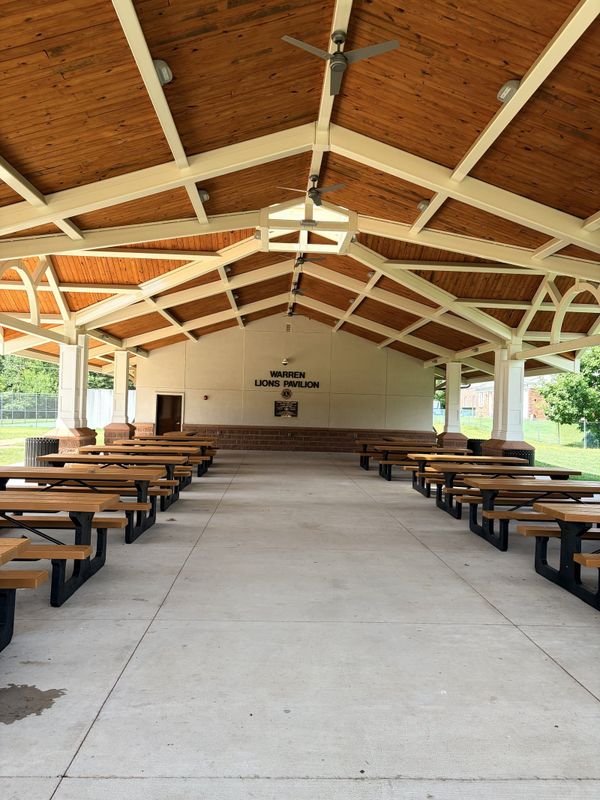 The Warren Lions Pavilion, an open air timber pavilion with a pitched wood ceiling and rows of picnic tables, WARREN LIONS PAVILION lettered on the end wall and the ball field fence beyond.