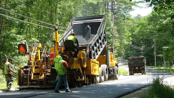 A road crew laying fresh asphalt behind a paver on a tree lined two lane road.