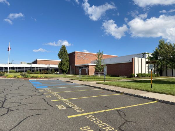 The Watchung Hills Regional High School campus, the lettered brick block and low window wings seen across the reserved parking lot, with the flagpole and flag at left.