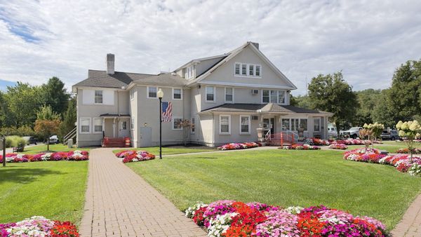 The Hofheimer House, a gray stucco house with a Welcome to Warren flag banner on a lamppost, set behind a brick walk and beds of red, pink and white impatiens.