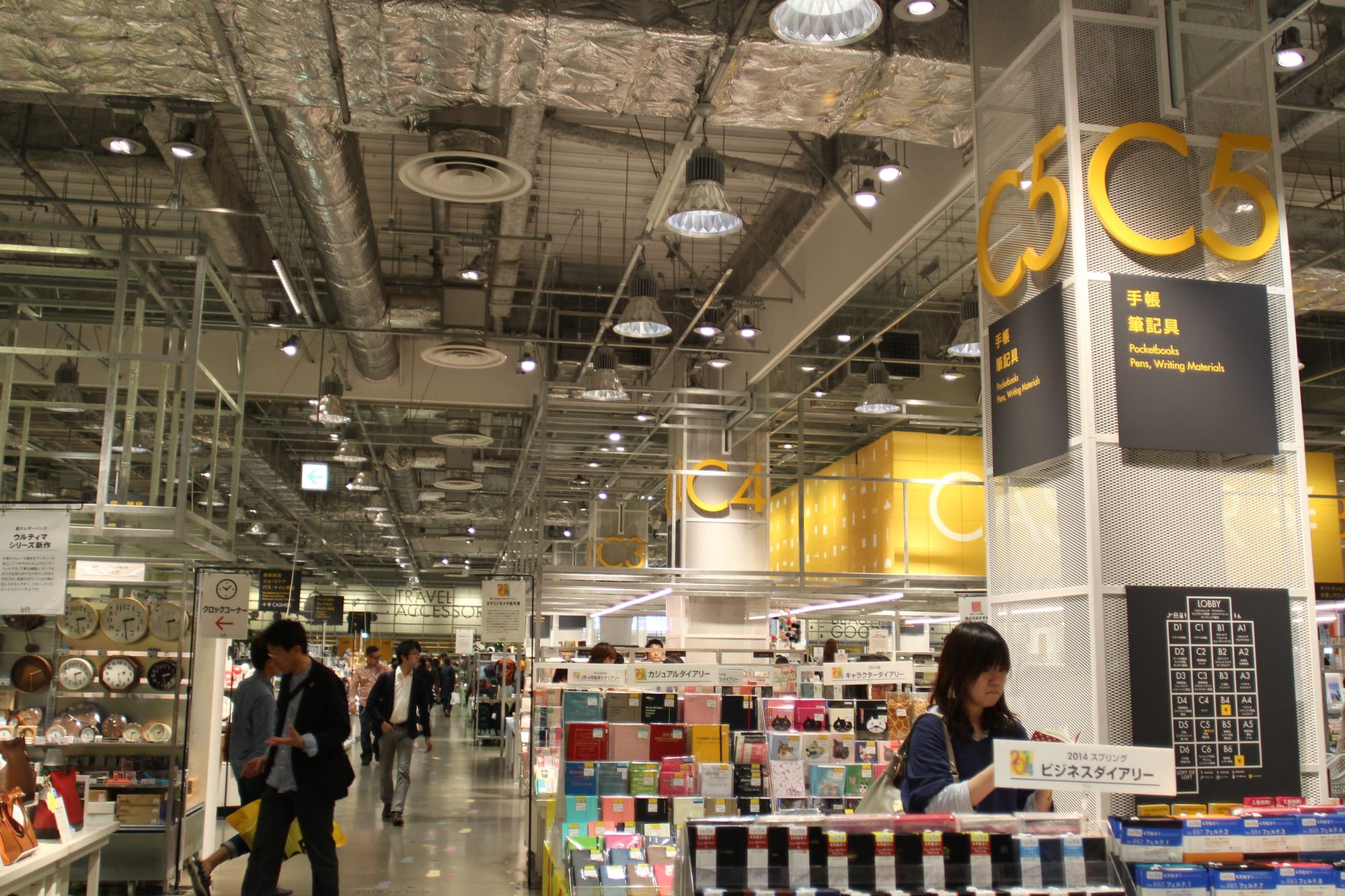 Shoppers browse the LOFT store. The brightly lit stationery section with yellow signs and exposed ceiling ducts.
