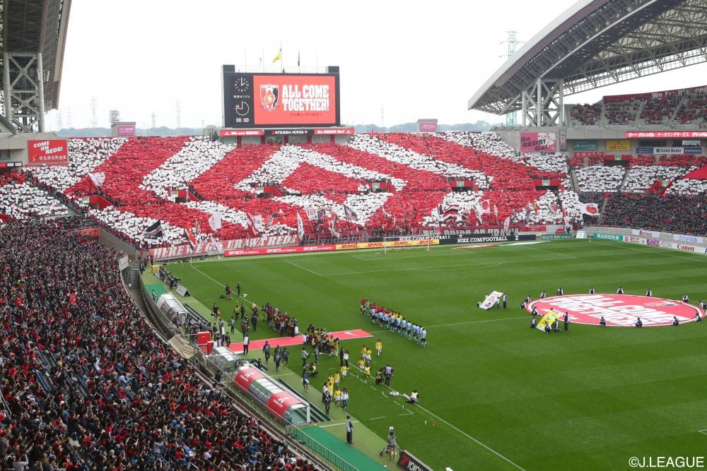 Stadium full of Urawa fans in red and white creating patterns, teams on the field.