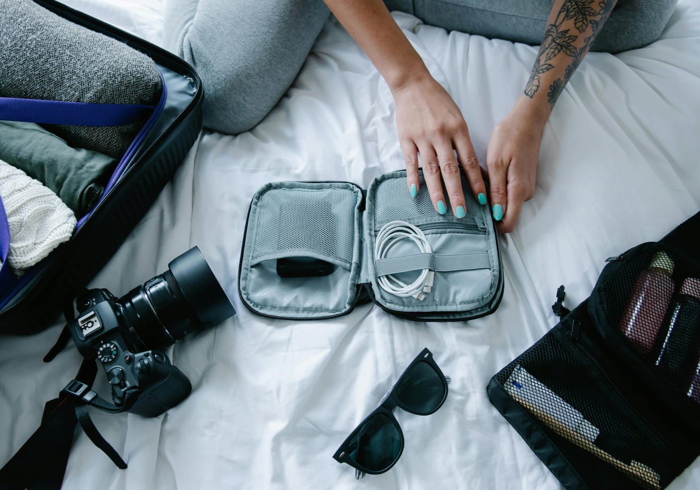 Person organizes cables in a pouch on a white bed. Nearby are a camera, open suitcase, sunglasses, and toiletry bag.