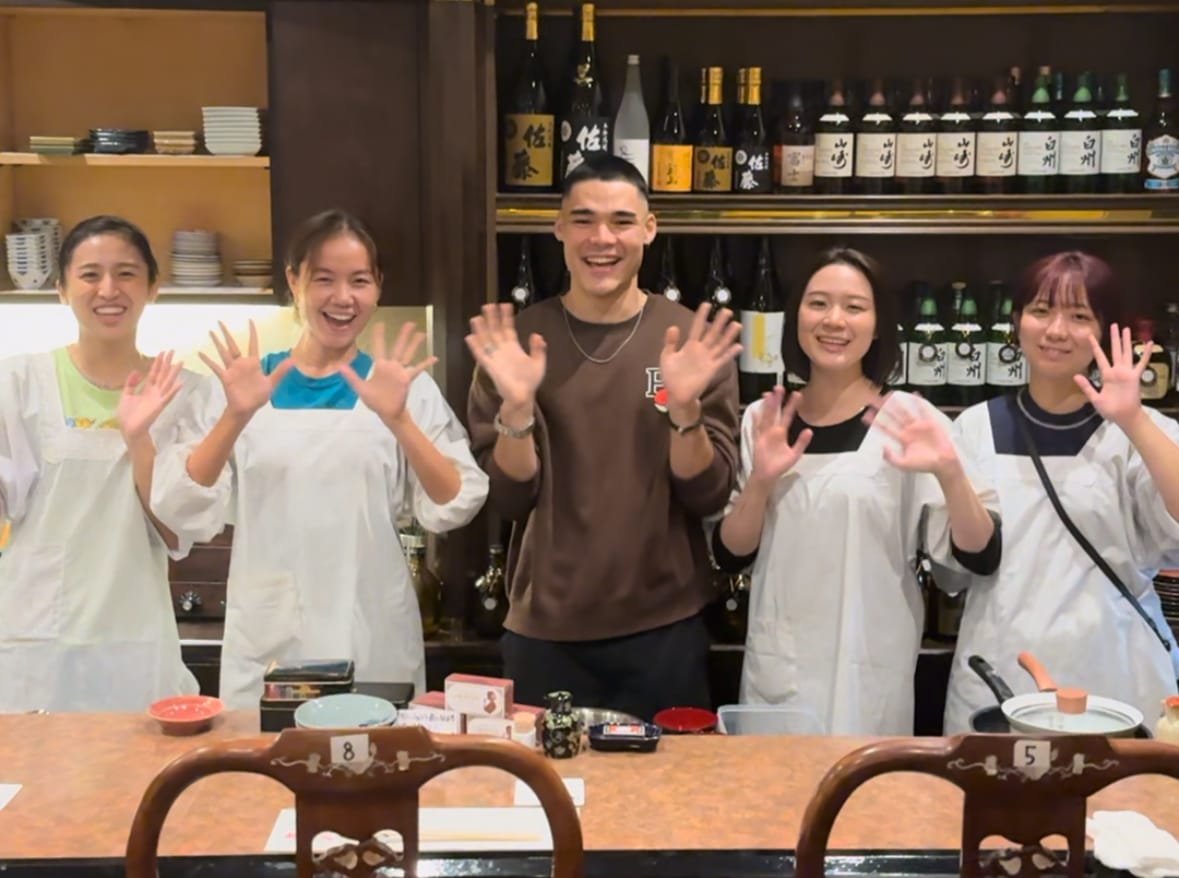 A young Japanese man at a local Japanese bar standing next to 4 Japanese waiters