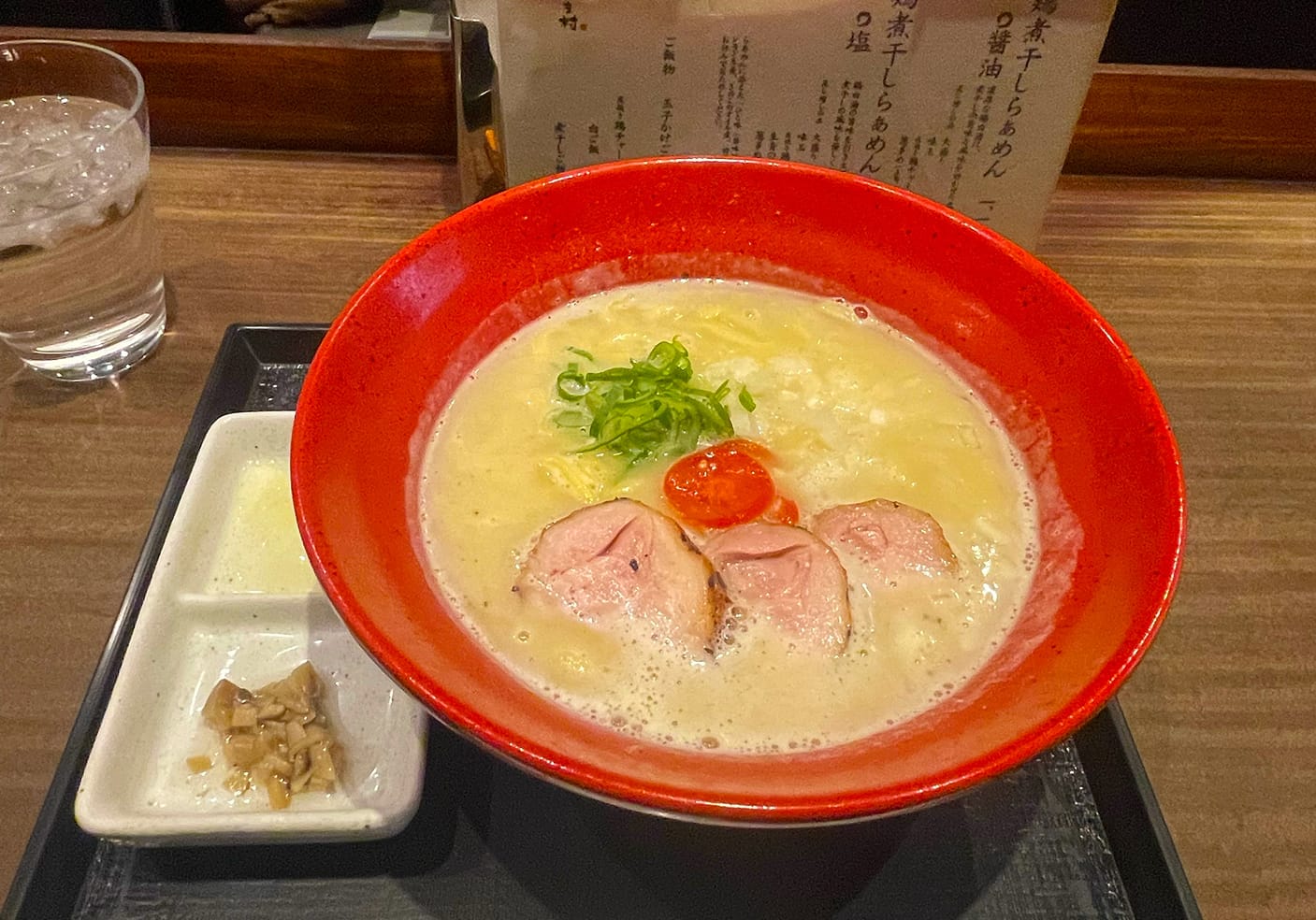 The signature chicken soup ramen at Menya Imamura. Red bowl of ramen with meat, scallions, and tomato.
