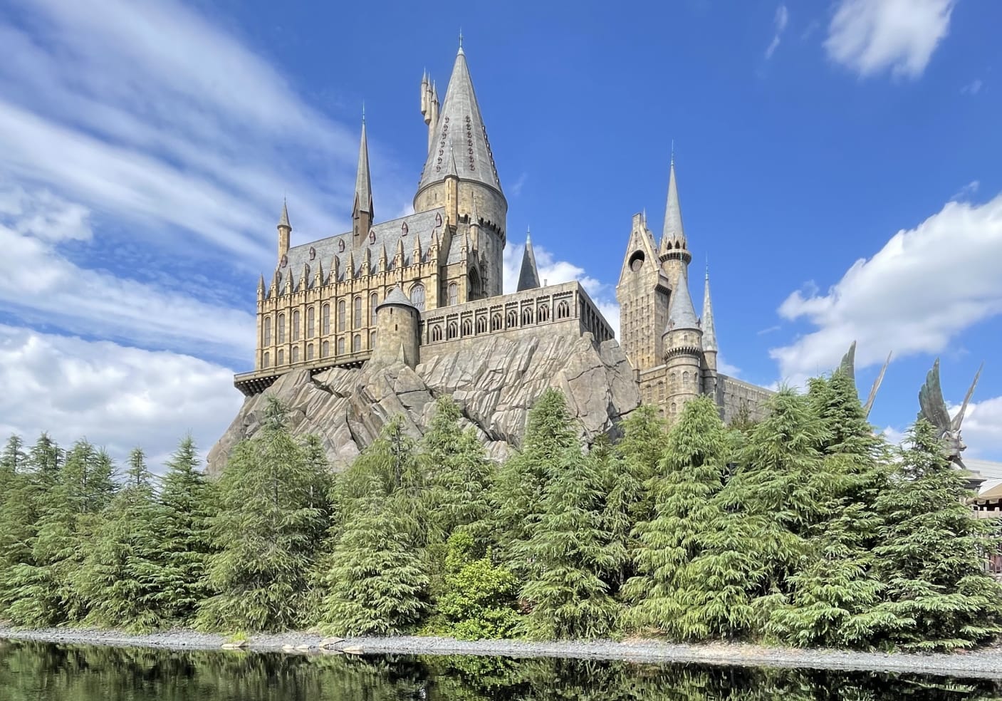 Majestic Hogwarts Castle surrounded by green trees, reflecting in the Black Lake under a bright blue sky with clouds.