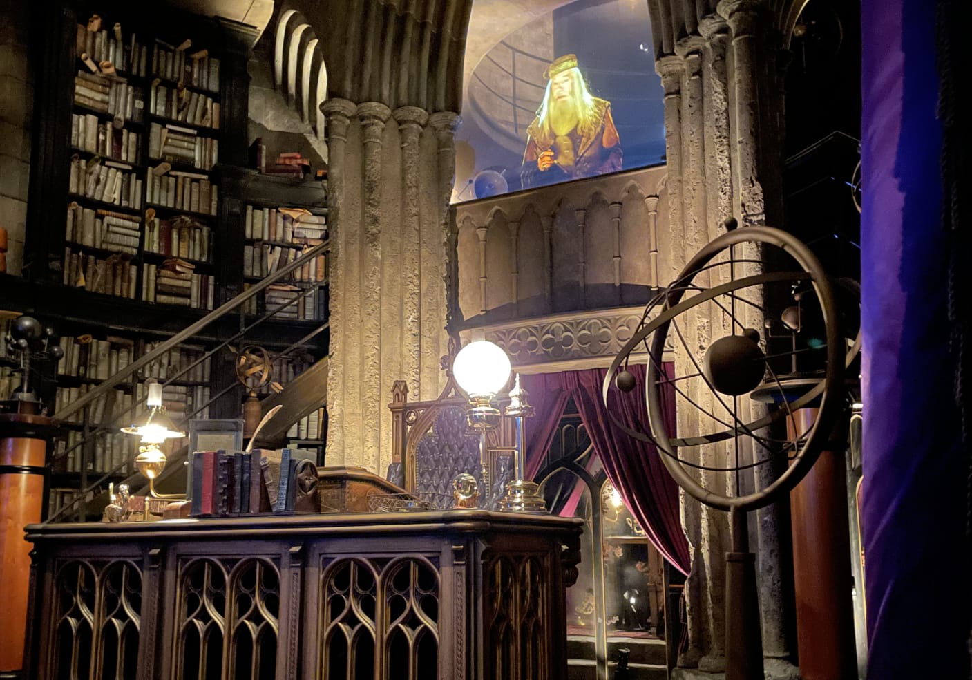 Dumbledore's office filled with bookshelves, globes, an ornate desk, purple drapes and stone arches. Dumbledore appears above on the upper level.