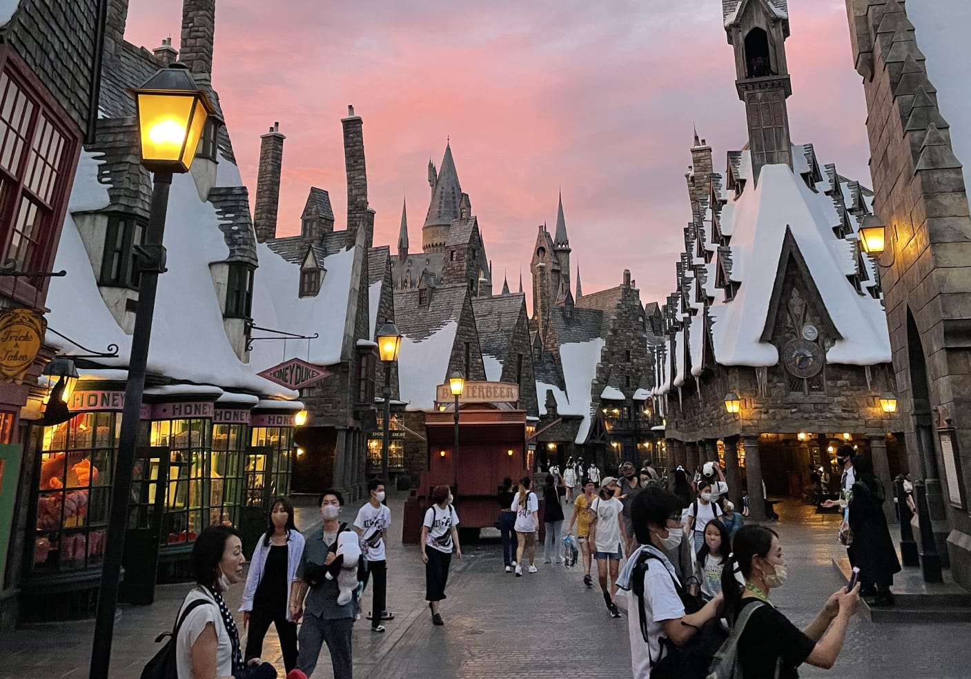 People walk through snowy Hogsmeade village at sunset, surrounded by shops with signs for Honeydukes and Butterbeer. 