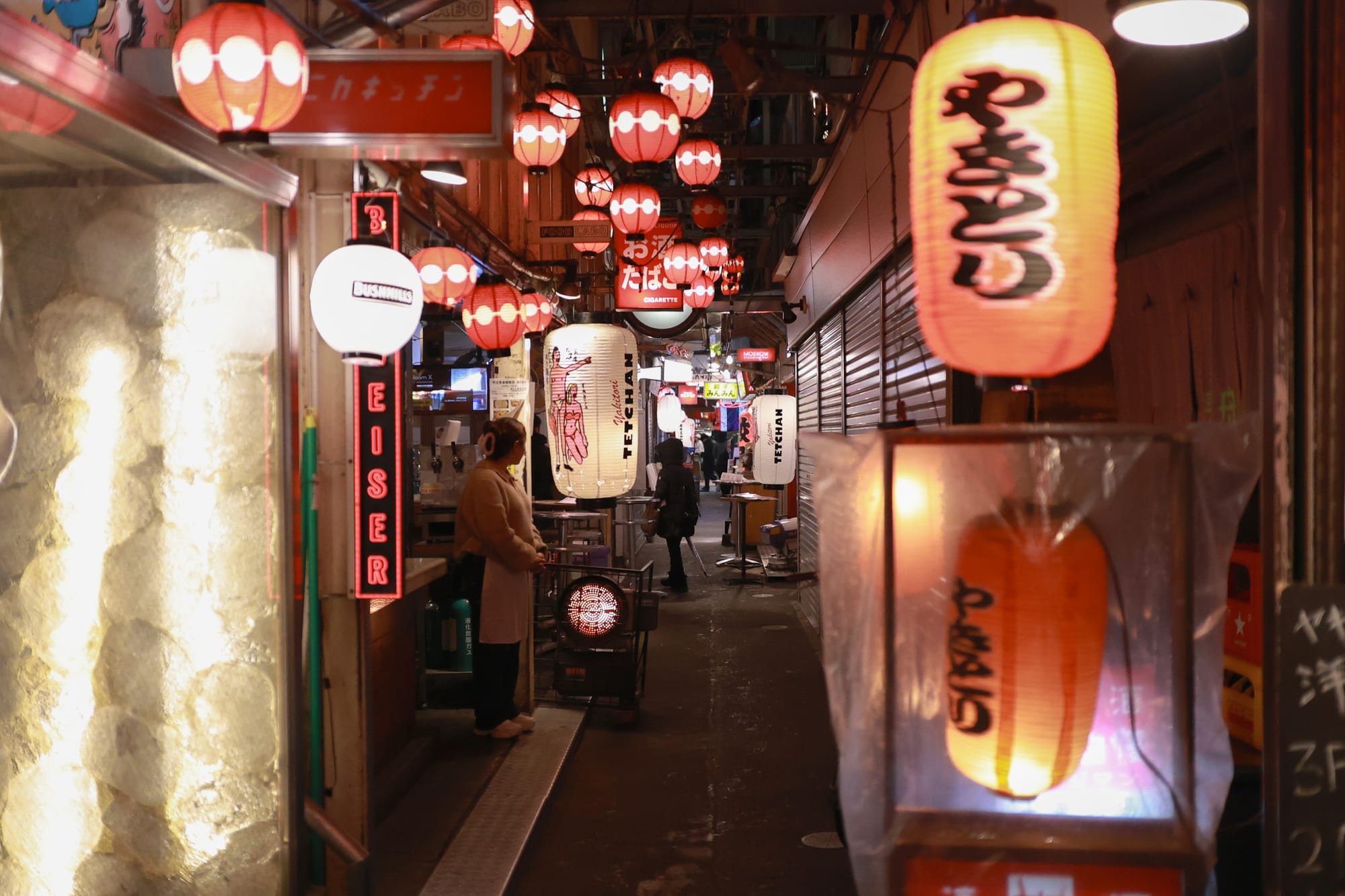 A street filled with bars at night in Kichijoji, with Japanese lanterns and some people outside.