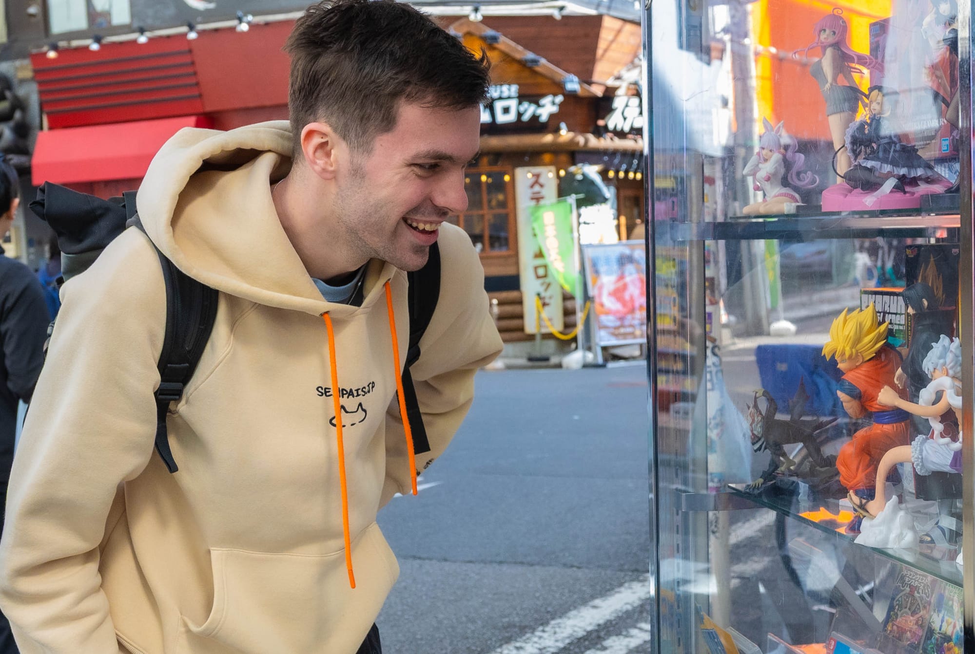 A young man in Japan looking at a shop that sells anime figures, waring a yellow hoodie and smiling.
